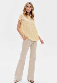 Jasnożółty sweter z dzianiny w diamentowy wzór, zestawiony z beżowymi spodniami dzwonowymi. Modelka ma na sobie brązowe sandały na obcasie.