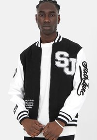 Jonge man met gevlochten haar in een zwart-witte varsityjack met de tekst "SJ" en "Sixth June", staande tegen een effen achtergrond.
