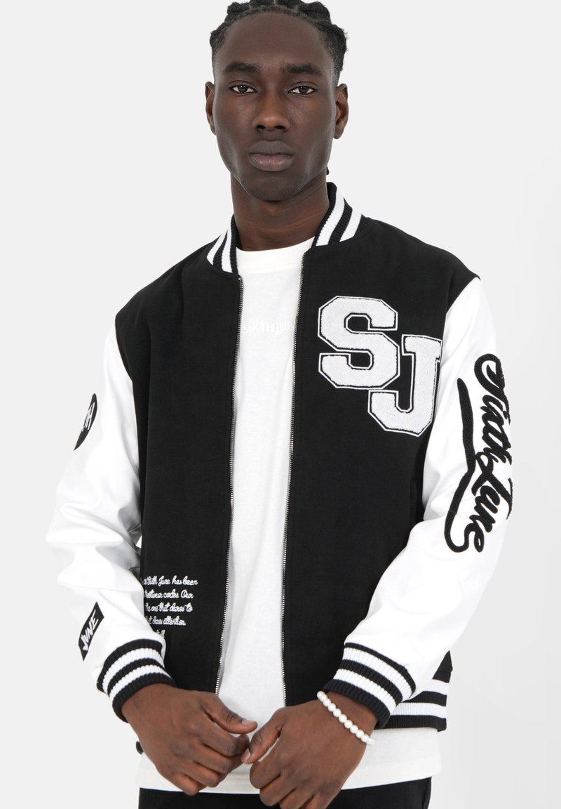 Jonge man met gevlochten haar in een zwart-witte varsityjack met de tekst "SJ" en "Sixth June", staande tegen een effen achtergrond.