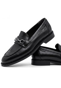 Paire de mocassins en cuir noir avec petits talons et détail chaîne argentée à l'avant, positionnée sur fond blanc.