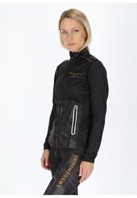 Schwarze Sportweste mit einem Reißverschluss vorne, ausgestattet mit einem strukturierten unteren Panel und reflektierenden Akzenten. Kombiniert mit gemusterten Leggings.