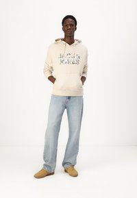 Haut à capuche beige avec poche avant et motif floral, associé à un jean bleu clair et des chaussures marron. Le modèle se tient avec les mains dans les poches.
