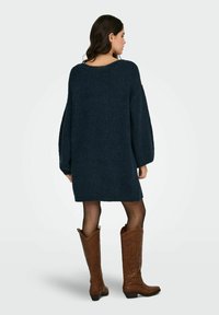 Granatowa, oversizeowa sukienka dzianinowa z długimi rękawami, fakturowaną tkaniną i szerokim dekoltem, zestawiona z brązowymi kowbojkami do kolan.