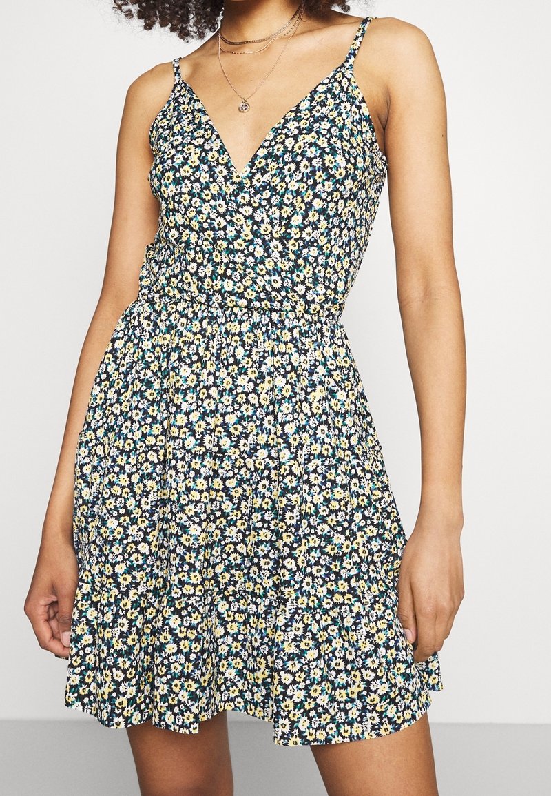 Femme portant une robe sans manches à imprimé floral avec des fleurs jaunes, blanches et bleues et un décolleté en V, accessoirisée de colliers superposés.