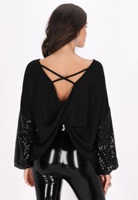 Maglione nero oversize con texture a coste, scollatura a V profonda sul retro, spalline incrociate e maniche decorate con paillettes. Abbinato a leggings neri lucidi.