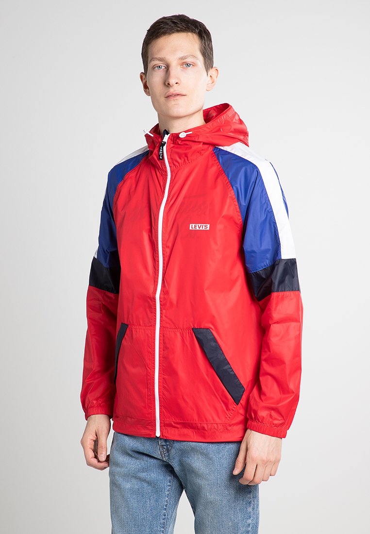 Veste légère rouge avec des accents bleus et blancs, fermeture éclair complète, capuche, deux poches latérales et poignets élastiques. Logo Levi's sur la poitrine.