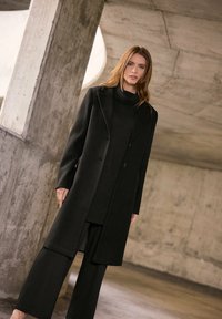 Cappotto lungo nero realizzato in tessuto testurizzato con colletto a risvolto, abbinato a un dolcevita e pantaloni a gamba larga, tutti di un nero uniforme.