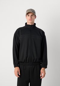 Casaco desportivo preto com fecho éclair, gola alta, punhos e barra elásticos. Apresenta costuras horizontais e um detalhe de logo subtil no peito.