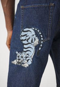 Jeans in denim blu scuro con un grande tigre ricamato blu e nero sulla tasca posteriore, che mette in mostra trame e motivi dettagliati.