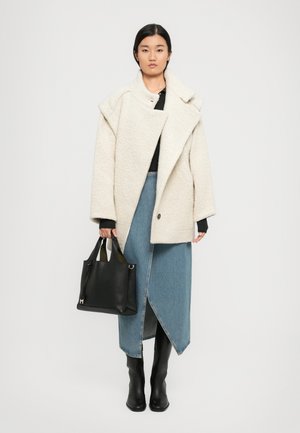 Palton oversized de culoare crem, cu o textură catifelată și guler lat, asortat cu o fustă din denim albastru și cizme negre până la genunchi, purtând o geantă din piele neagră.