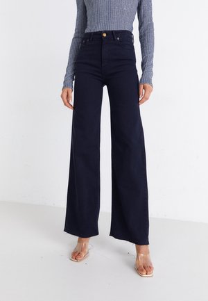 Persona vistiendo jeans negros de cintura alta y pierna ancha con dobladillo sin acabado y sandalias de tacón translúcidas con punta cuadrada, sobre un fondo blanco liso.