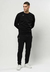 Bărbat purtând hanorac negru și pantaloni de trening cu mici logo-uri albe, stând cu mâinile la spate, privirea îndreptată spre dreapta, pantofi sport albi.