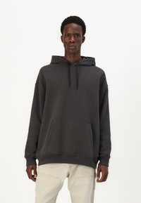 Tumši pelēks hoody, izgatavots no mīksta kokvilnas maisījuma, ar priekšējo kabatu, aukliņu kapuci un brīvu siluetu. Kombinēts ar gaišiem biksēm.