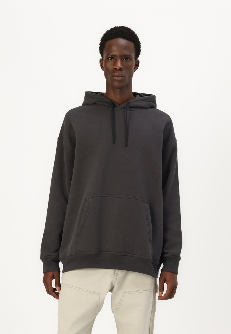 Tumši pelēks hoody, izgatavots no mīksta kokvilnas maisījuma, ar priekšējo kabatu, aukliņu kapuci un brīvu siluetu. Kombinēts ar gaišiem biksēm.