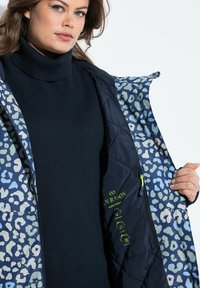 Námořnická prošívaná bunda s podšívkou v modro-zeleném leopardím vzoru, obsahující zipy a zelené logo; texturovaná látka a útulný rolák pod ní.