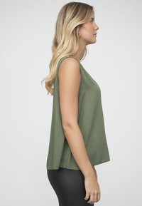 Olivetgrønn ermeløs topp med løs passform, rund hals og mykt stoff, kombinert med tettsittende svarte bukser. Ingen synlige mønstre eller metallbeslag.