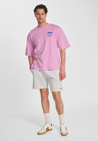 Rosa t-shirt med kort ärm och blå grafisk tryck, tillsammans med ljusgrå shorts och vita sneakers med svarta detaljer.