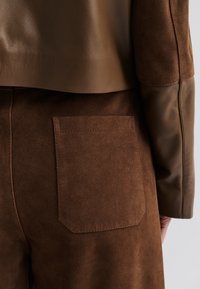 Pantaloni in suede marrone con top in pelle, caratterizzati da una tasca posteriore quadrata e trame a contrasto. Le maniche in pelle liscia esaltano il design.