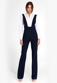 Kobieta stoi w granatowych spodni wysokiej talii z szerokimi szelkami, nosząc białą koszulę zapinaną na guziki i beżowe obcasy na białym tle.