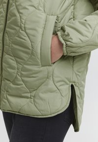 Pikowana zielona kurtka z bocznymi kieszeniami, elastycznymi ściągaczami przy rękawach i lekko zaokrąglonym dołem. Teksturowana powierzchnia z widocznymi detalami przeszyć.