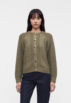 Kvinde med lige mørkt hår iført olivengrøn kabelstrikket cardigan med store knapper og mørke jeans, stående foran ensfarvet baggrund.