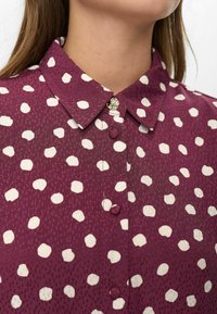 Gros plan sur une chemise bourgogne à pois blancs irréguliers, avec des boutons recouverts de tissu et un bouton de col supérieur décoratif en or.
