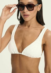 White bikini top with triangle cups featuring textured fabric, wide straps, and a gathered band. Accompanied by oversized dark sunglasses.