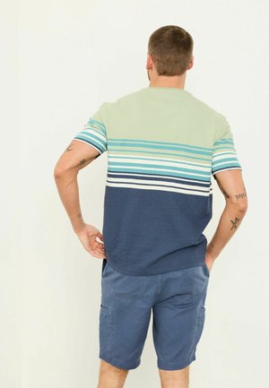 Bărbat cu păr scurt, stând cu mâinile pe șolduri, purtând o cămașă cu mâneci scurte în dungi și pantaloni scurți cargo albaștri, cu spatele către cameră.