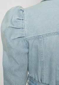 Svetlo modra jeans jakna z napihnjenimi rameni, surovimi robovi in pasom z obićajnim ovratnikom.