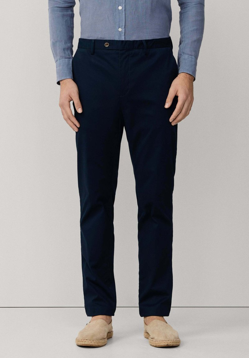 Man draagt marineblauwe broek, lichtblauw overhemdbloesje en beige instapschoenen, staand tegen een effen lichte achtergrond.