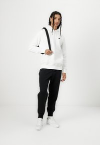 Felpa bianca con tasca a marsupio, abbinata a pantaloni jogger neri. Presenta una cinghia nera sulla spalla, completata da sneakers bianche.