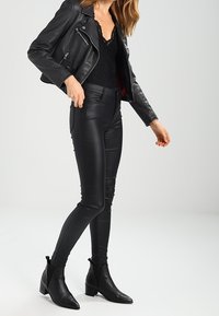 Veste en cuir noir avec fermetures éclair, haut en dentelle noire et pantalon noir ajusté. Bottes à hauteur de cheville avec un bout pointu et un talon carré.