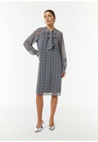 Blumendruck, knielanges Kleid aus leichtem, navyfarbenem Stoff mit einem Schleifenkragen und langen, transparenten Ärmeln. Getragen mit silbernen High Heels.