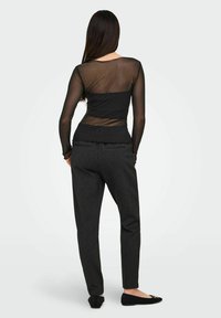 Kobieta z długimi włosami, ubrana w czarną przezroczystą bluzkę z długim rękawem, szare spodnie w prążki oraz czarne baleriny, stojąca tyłem na białym tle.