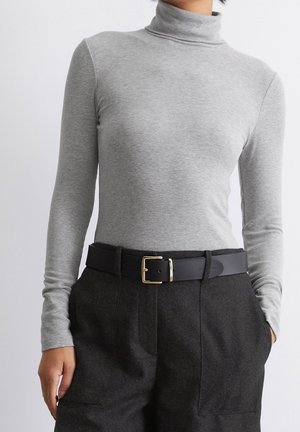 Personne portant un pull à col roulé gris clair rentré dans un pantalon taille haute gris foncé avec une ceinture noire et une boucle dorée.