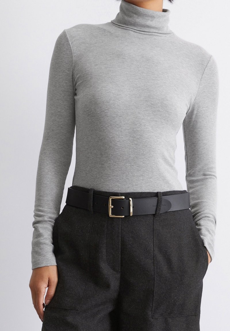 Personne portant un pull à col roulé gris clair rentré dans un pantalon taille haute gris foncé avec une ceinture noire et une boucle dorée.