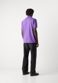 Μωβ πόλο μπλούζα με κοντά μανίκια, υφασμάτινη υφή και μαύρες λεπτομέρειες στους ώμους, συνδυασμένη με σκούρους παντελόνες και μαύρα παπούτσια.