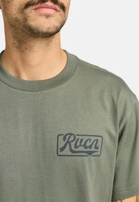 Olivegrünes Baumwoll-T-Shirt mit Rundhalsausschnitt, das auf der linken Seite mit einem dunkelgrauen "Rvca"-Logo bedruckt ist. Glatte Textur, kurze Ärmel.