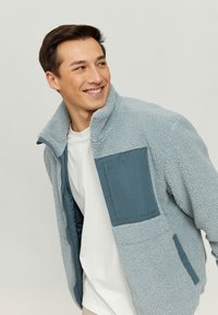 Fleece jacket in light blue with a dark blue chest pocket, zip closure, and high collar; soft texture and casual design.
