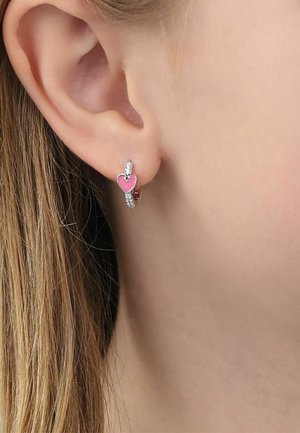 Pendiente de aro plateado con pequeño charm en forma de corazón rosa, llevado en el lóbulo de la oreja de una persona de piel clara y cabello rubio.