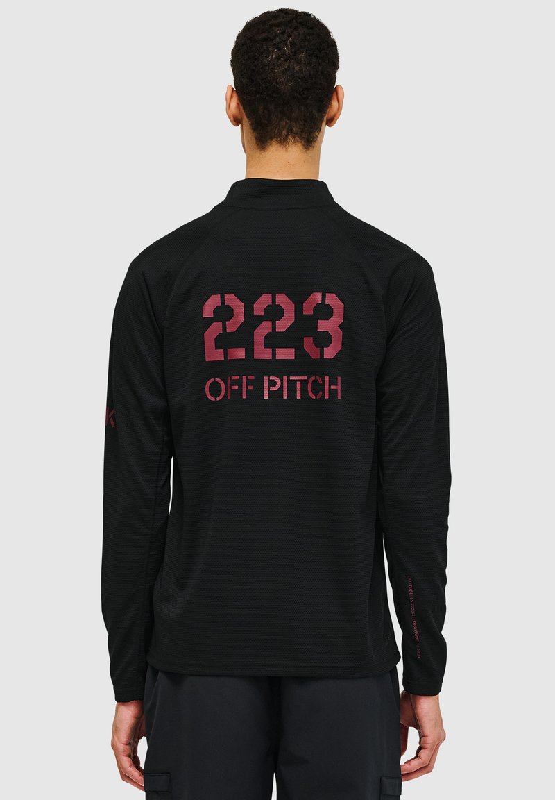 Chemise noire à manches longues en tissu texturé. Présente de grands chiffres rouges "223" et "OFF PITCH" imprimés dans le dos, avec un col rond.