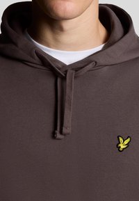 Brun hættejakke med snørre, der har et gult broderet logo af en fugl på brystet. Blødt stof, afslappet design.