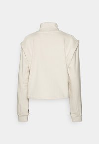 Sudadera recortada de color crema con un cuello alto, mangas largas y una textura suave. Presenta aberturas laterales en los hombros.