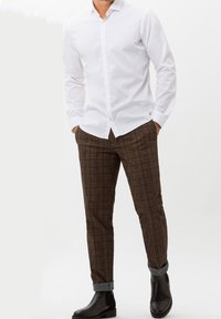 Homme les mains dans les poches portant une chemise blanche boutonnée, un pantalon marron à carreaux avec des revers, et des bottines Chelsea en cuir noir, debout sur un fond blanc.