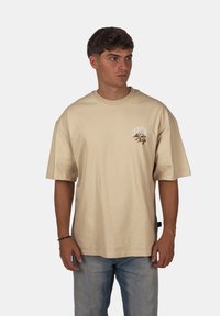 Béžové bavlněné tričko s krátkým rukávem, kulatým výstřihem a malou grafickou nálepkou s nápisem "Crystal" na levé části hrudi.