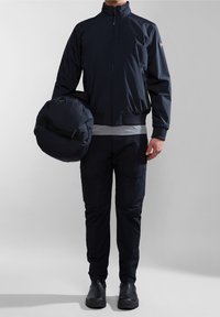 Marineblå bomberjakke med lynlås, ribbede manchetter og talje, parret med matchende cargo-bukser. Modellen holder en rund, polstret taske.