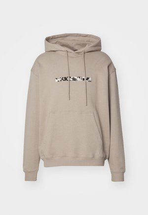 Beige hettegenser med en stor frontlomme, snorhette og et svart-hvitt mønstret logo på brystet. Myk bomullsstoff, uformell design.