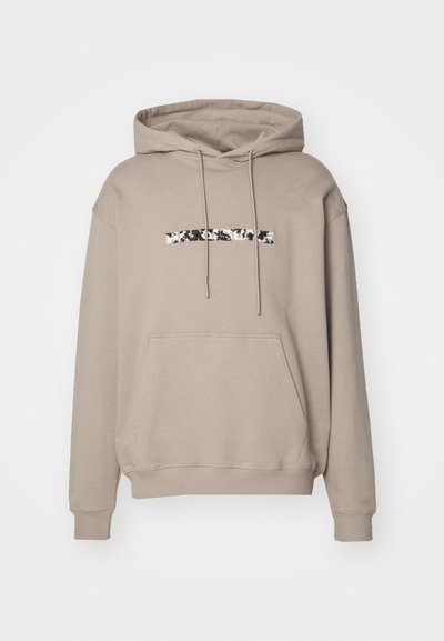 Sudadera beige con un gran bolsillo delantero, capucha con cordón y un logo de patrón en blanco y negro en el pecho. Tela de algodón suave, diseño casual.