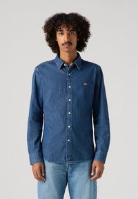 Sötétkék farmering gombos elülső résszel, hegyes gallérral, hosszú ujjakkal és egy kis piros Levi’s logóval a mellkasán. Simább textúrával.