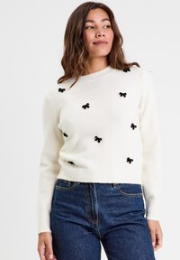 Pull en tricot blanc avec des accents en tissu noir en forme de nœud, des poignets et un ourlet côtelés. La coupe est ajustée avec un col rond et une texture douce.